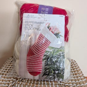 May include: A clear plastic bag containing red and white yarn skeins and a printed pattern insert labeled Annie's Holiday Crochet Pattern Kit Club Peppermint Stick Stocking, featuring a photo of a crocheted red and white striped stocking.
