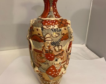 Puede incluir: Un jarrón ornamentado con una parte superior acampanada y un diseño detallado pintado a mano. El jarrón presenta una base de color crema, un cuello rojo y naranja y un cuerpo adornado con figuras con vestimenta tradicional. La estética general recuerda al arte japonés.