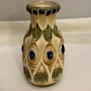 Könnte beinhalten: Kleine, dekorative Keramikvase mit cremefarbenem Grund und aufwendigen Details. Die Vase weist ein Muster aus grünen Blättern, goldenen Akzenten und blauen kreisförmigen Verzierungen auf. Der Rand ist mit kleinen blauen Punkten verziert.