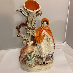 May include: A ceramic vase with a tree trunk design and a hollowed-out top. A figure in a red hooded cloak and white dress sits beside the tree, holding a basket. A dog is at the base of the tree. The vase has a cream-coloured base and is painted in various colours.