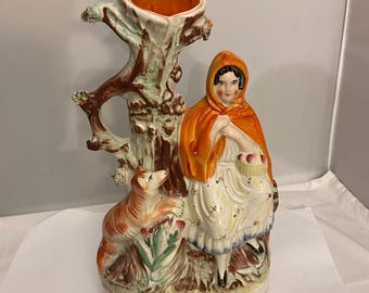 May include: A ceramic vase featuring a tree trunk design with a hollowed-out top. A figure in a red hooded cloak and white dress sits beside the tree, holding a basket. A dog is at the base of the tree. The vase has a cream-colored base and is painted in various colors.