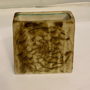 Puede incluir: Un jarrón cuadrado de cerámica con un exterior marrón texturizado y un borde verde claro. El jarrón presenta un patrón en relieve detallado en sus lados, agregando un elemento decorativo único. El interior es de color beige claro.