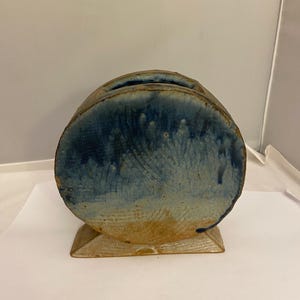 May include: A handcrafted ceramic vase with a unique, rounded shape and a rectangular base. The vase features a striking blue glaze that drips down the sides, contrasting with the tan base. A slot is cut into the top.