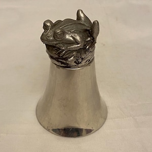Pode incluir: Um saleiro prateado com uma figura de rã no topo. A rã tem pele texturizada e assenta sobre uma faixa decorativa. O saleiro tem uma base alargada e um acabamento liso e polido.