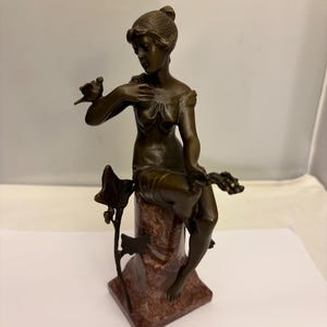 May include: Bronze sculpture of a seated woman with a bird on her hand. The figure is atop a marble base with a square bottom. The sculpture has a dark brown patina and is detailed with flowing lines and curves.