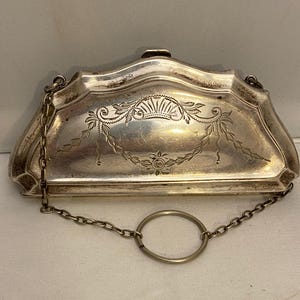 May include: An antique silver-coloured purse with a decorative floral and leaf design. It has a chain handle and a circular ring. The clasp is at the top. The purse is rectangular with curved edges.