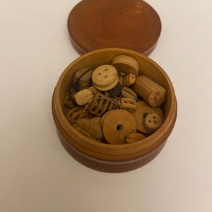 May include: A small, round wooden box with a lid, filled with various wooden buttons and small wooden pieces. The buttons are of different sizes and shapes, some with holes and others with decorative details. The box and lid are a warm brown colour.