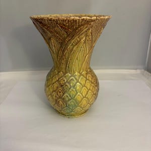 May include: A ceramic vase with a pineapple-inspired design, featuring a textured, diamond-patterned body and a flared, leaf-like rim. The glaze is a blend of yellow, green, and brown tones, creating a weathered, multi-coloured appearance.