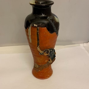 May include: A ceramic vase with a dark brown top and a reddish-orange body. The vase features a sculpted figure of a person climbing a tree branch. The vase is approximately 10 inches tall.