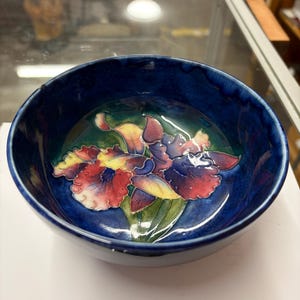 Puede incluir: Un cuenco de cerámica redondo con un exterior e interior esmaltados en azul intenso. El centro del cuenco presenta un diseño floral en relieve y colorido que representa orquídeas en tonos rojos, morados, amarillos y verdes sobre un fondo azul oscuro.
