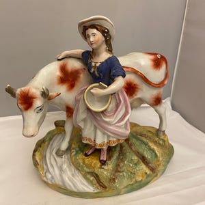 Puede incluir: Una figurita de porcelana de una mujer con un vestido azul y un sombrero blanco, sosteniendo un cubo, de pie junto a una vaca blanca con manchas marrones.