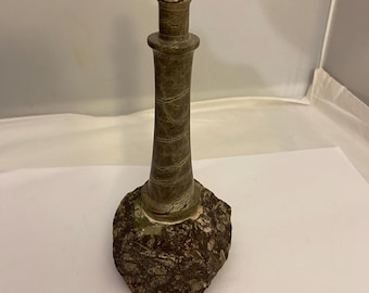 Puede incluir: Una escultura decorativa de un faro. El faro es de color verde-marrón apagado con una superficie texturizada y se asienta sobre una base de piedra rugosa de color marrón oscuro. El faro tiene un cuerpo cilíndrico y una pequeña parte superior redondeada.