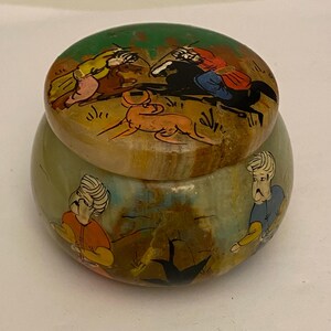 Könnte beinhalten: Eine runde, dekorative Dose mit Deckel und handgemalten Illustrationen. Der Deckel zeigt Figuren auf Pferden und einen Hund vor grünem Hintergrund. Der Körper der Dose hat ähnliche Figuren in Orange und Blau.