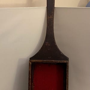 May include: A dark brown wooden box with a long handle. The box has a rectangular shape with a red felt interior. The handle is long and slender, and the box appears to be handmade.