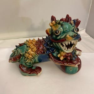 Può includere: Statuetta di drago in ceramica nei toni del verde, blu, rosso e giallo. Il drago ha denti affilati, una criniera dettagliata ed è posato su una base rotonda. La statuetta è un oggetto decorativo.