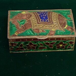 May include: A small rectangular box with an intricate design. The box is made of metal and has a green background with a red and gold elephant design. The lid of the box is decorated with a floral pattern in red, green, and gold.