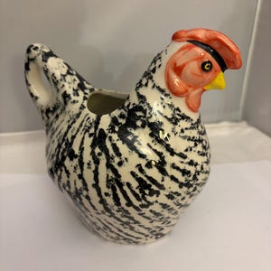 May include: A ceramic chicken-shaped creamer with a black and white speckled body. The head features a red comb, a yellow beak, and a black eye. The creamer has a handle and a spout for pouring.