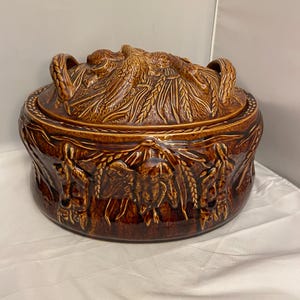 Puede incluir: Una fuente de cerámica marrón con tapa. La tapa presenta un diseño de espiga de trigo y dos asas. La fuente tiene forma redonda y está decorada con relieves de trigo y otros elementos. La fuente es probablemente vintage.