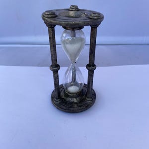 Può includere: Una clessidra in stile antico con finitura bronzo metallizzato. La lampadina di vetro contiene sabbia bianca, tenuta tra due piastre circolari collegate da quattro supporti verticali. La clessidra è un oggetto decorativo.