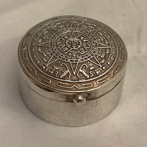 Puede incluir: Caja de alhajas de plata con una tapa circular que presenta un diseño intrincado del calendario azteca. La caja está hecha de plata pulida y tiene una forma redonda y lisa.