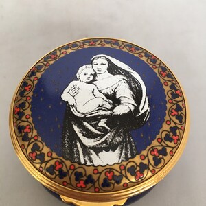 May include: A round, blue and gold trinket box with a white image of the Virgin Mary holding a baby Jesus. The box is decorated with a gold border and red and gold floral designs.