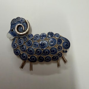 May include: A silver-toned brooch shaped like a ram, featuring intricate blue enamel detailing on its curled fleece and head. The design incorporates metallic silver filigree swirls throughout the body, with a smooth white accent on the nose.