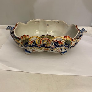 Może przedstawiać: Ozdobna ceramiczna miska z falistym brzegiem i trzema małymi nóżkami. Miska ma białe wnętrze i jest ozdobiona kolorowymi wzorami kwiatowymi i liściastymi w kolorze czerwonym, niebieskim, żółtym i zielonym. Ma dwa ozdobne uchwyty po bokach.