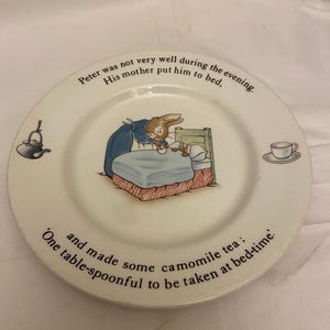 May include: A white ceramic plate featuring Peter Rabbit in bed. The plate has the text "Peter was not very well during the evening. His mother put him to bed. and made some camomile tea: One table-spoonful to be taken at bed-time."