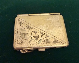 May include: A rectangular, antique gold-colored metal box with a hinged lid. The lid features an engraved design of swirling patterns and lines. The box has a small loop on the side and a clasp to secure it closed. The box is on a dark green surface.