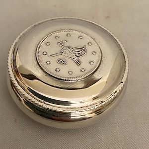May include: A silver round box with a raised circular design on the lid. The design features a bird with a starburst around it.