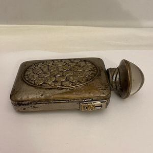 Peut inclure: Une flasque en métal vintage avec un motif texturé de forme ovale sur le couvercle. La flasque a une petite ampoule en verre transparente et ronde à une extrémité et un petit loquet sur le côté. Le métal a un aspect antique terni.