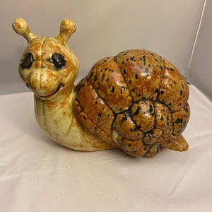 May include: A ceramic snail figurine with a brown and tan shell with black spots. The snail has a light brown body with black eyes and a smiling face.