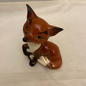 Könnte beinhalten: Eine Keramikfigur eines Fuchses, der mit seinen Pfoten unter dem Körper sitzt. Der Fuchs ist braun mit schwarzen Markierungen und weißen Akzenten.