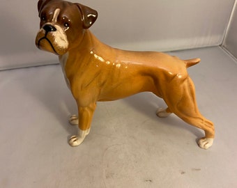 May include: A ceramic figurine of a brown and white boxer dog. The dog is standing with its head turned to the side and its tail curled.