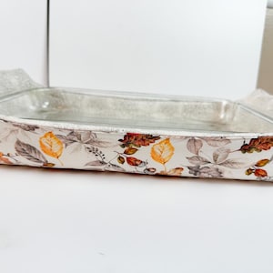 Puede incluir: Un plato para hornear blanco con un patrón floral que presenta hojas y bellotas. El plato está hecho de vidrio y tiene una cubierta de tela blanca con un patrón floral.