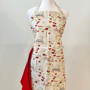 May include: A cream-colored apron with a red and white cherry and heart pattern. The apron has two pockets and the text "Kiss the Cook" and "Bless the Baker" is printed on the apron.