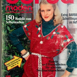 May include: A magazine cover featuring a woman wearing a red sweater with white and green embroidery. The magazine is titled "Burda Moden" and the text "150 Modelle zum Selbermachen" is visible. The cover also includes the text "Extra: BARBIE Schnittbogen im Hef" and "Basteln und Handarbeiten für schönste Weihnachtsgeschenke".