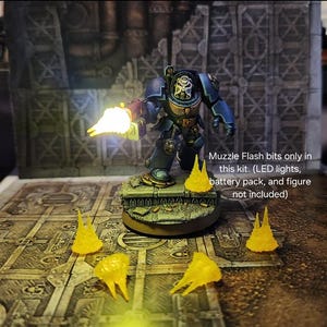 Puede incluir: Una miniatura de Space Marine azul con un efecto de destello de boca amarillo. La miniatura está de pie sobre una base con una textura verde y marrón. La imagen también muestra varios efectos de destello de boca amarillos sobre una superficie texturizada marrón. Los bits de destello de boca solo están disponibles en este kit. (Luces LED, paquete de baterías y figura no incluidos)