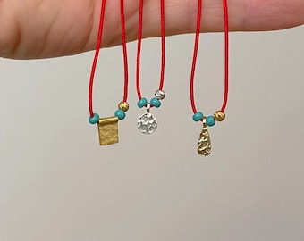 May include: Three delicate necklaces with red cords. Each necklace features a different pendant: a textured gold rectangle, a hammered silver disc, and a textured gold teardrop. Turquoise beads accent each pendant.