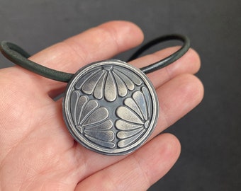May include: A silver pendant necklace with a circular design featuring a stylised floral pattern. The pendant is attached to a black leather cord.