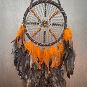 Peut inclure: Un attrape-rêves noir et orange avec un motif de toile d'araignée et une araignée noire au centre. L'attrape-rêves a des plumes noires et orange qui pendent du bas.