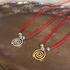 Op de afbeelding: Twee rode koordkettingen met turquoise kralen en een zilveren en gouden spiraal hanger. De kettingen liggen op een zilveren dienblad.