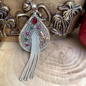 Könnte beinhalten: Eine Halskette mit einem tropfenförmigen Anhänger aus Silber und Roségold. Der Anhänger ist mit filigranen Verzierungen versehen und mit roten und grünen Edelsteinen besetzt. Die Halskette hat eine lange silberne Kette mit einem Quastenanhänger.