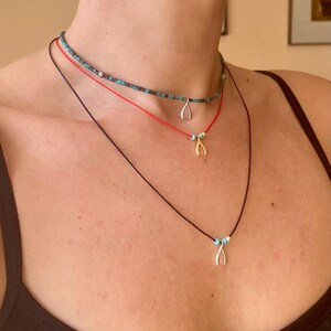 Può includere: Tre collane con perline turchesi e un ciondolo a forma di osso della fortuna. La collana superiore è un choker nero con perline turchesi. La collana di mezzo è rossa con perline turchesi e un ciondolo a forma di osso della fortuna dorato. La collana inferiore è nera con perline turchesi e un ciondolo a forma di osso della fortuna argentato.