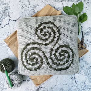 May include: A grey crocheted cushion with a green Celtic knot design. The cushion is on a brown paper background.