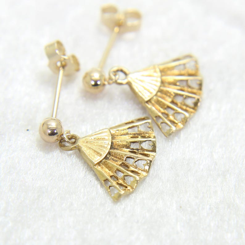 May include: A pair of gold-colored earrings featuring a fan design. Each earring has a post and a small ball, with a fan-shaped pendant. The fan pendants have a detailed, openwork design.