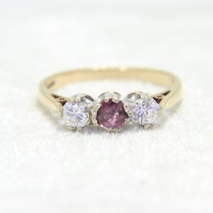 May include: A gold ring with three gemstones. The center stone is a pink gemstone, flanked by two clear, round-cut gemstones. The ring has a classic, elegant design, suitable for engagement or special occasions.
