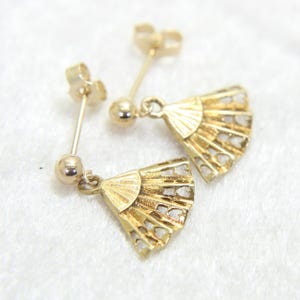 May include: A pair of gold-colored fan-shaped earrings. Each earring features a post with a small ball and a fan pendant with heart-shaped cutouts. The earrings are displayed on a white surface.