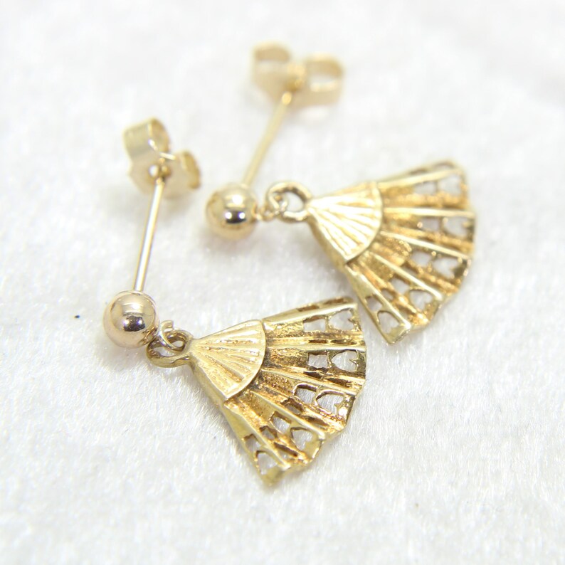 May include: A pair of gold-colored fan-shaped earrings. Each earring features a post with a small ball and a fan pendant with heart-shaped cutouts. The earrings are displayed on a white surface.