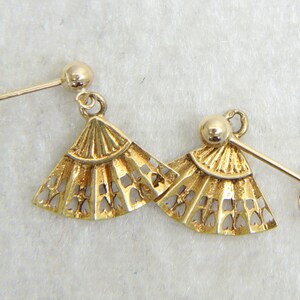 May include: A pair of gold-colored fan-shaped earrings. Each earring features a detailed, openwork design with a post and a small ball at the top. The earrings are displayed on a light-colored surface.